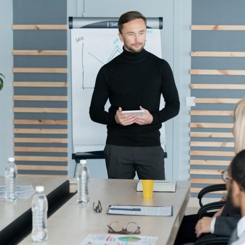 Content handsome young company leader with tablet standing at conference table and looking at colleagues while running meeting in office